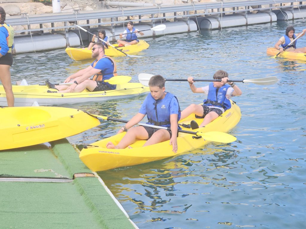 20260707_1708263318455241813437881 Group of children in yellow kayaks wearing blue life jackets, paddling near a dock during a boat lesson on calm blue water.