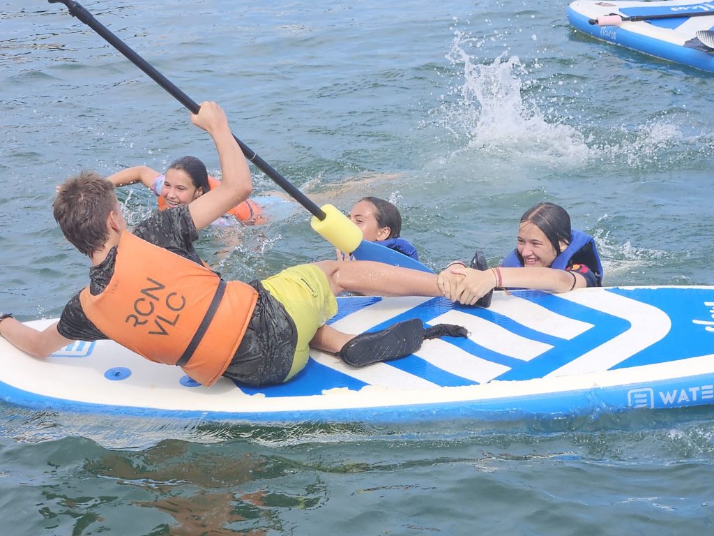 20260707_1712595599604687862736627 Group of kids and a teen on a blue-and-white paddleboard in the water, wearing orange life jackets and holding a paddle together.