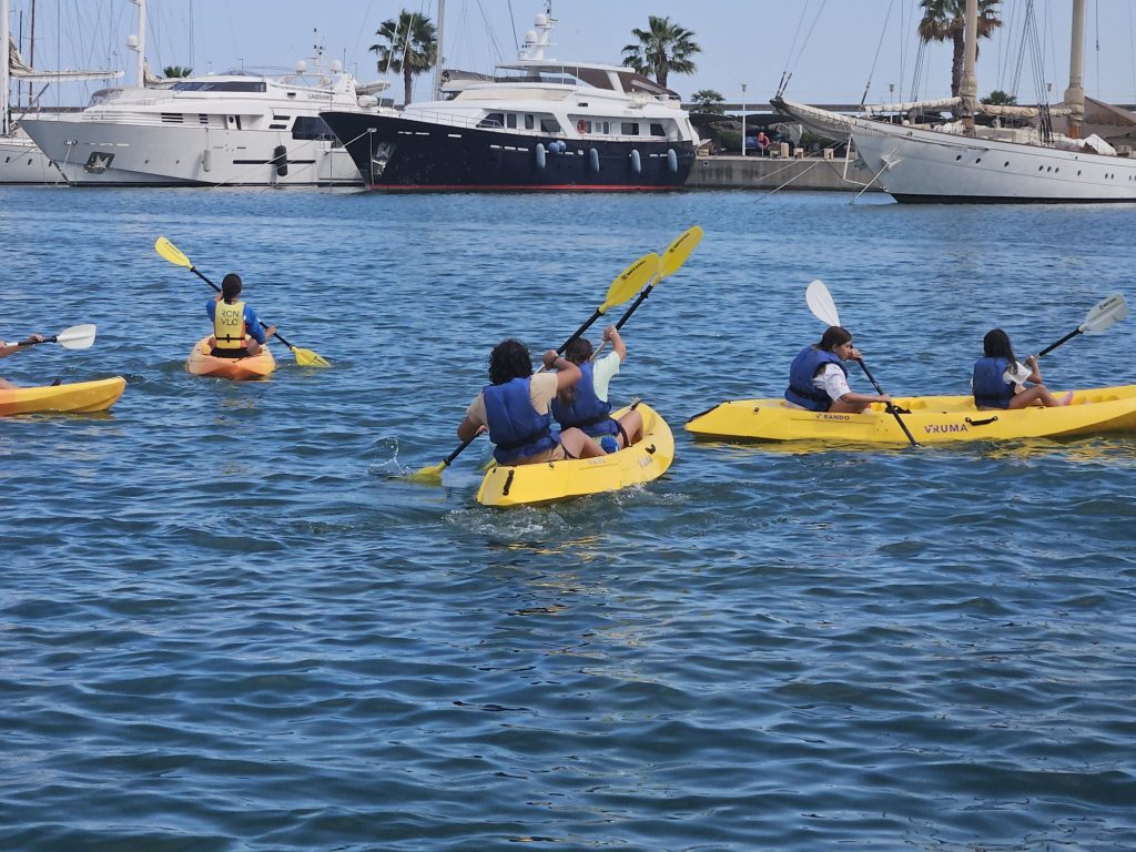 20260707_1713434664226952770833271 People in bright yellow kayaks wearing life jackets paddle in a sunny marina, with large luxury yachts moored along a quay in the background.
