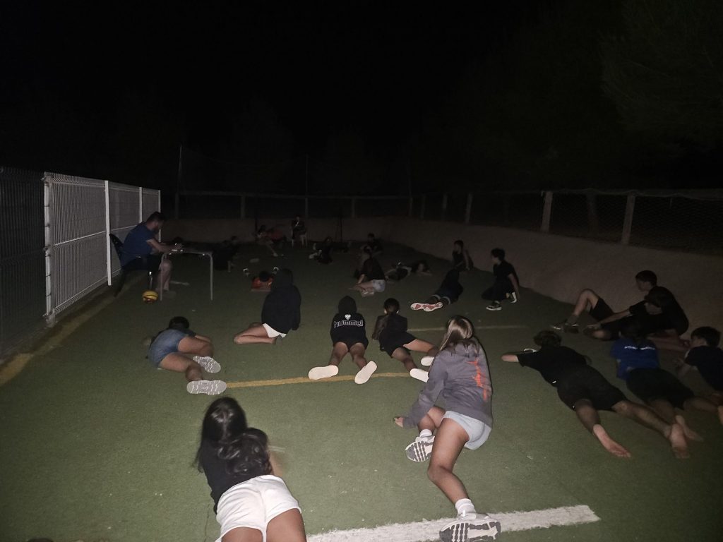 20260707_2255296680708210448990948 Group of kids lying and sitting on a nighttime sports field beside a chain-link fence, dimly lit.