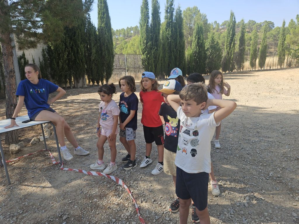 Group of children lined up outdoors behind a red-and-white barrier tape, waiting on a sunny dirt lot with trees in the background.