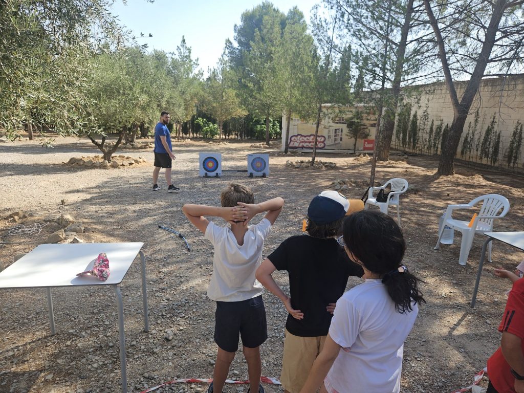 Children observe an archery lesson outdoors; a man in a blue shirt stands near two archery targets.