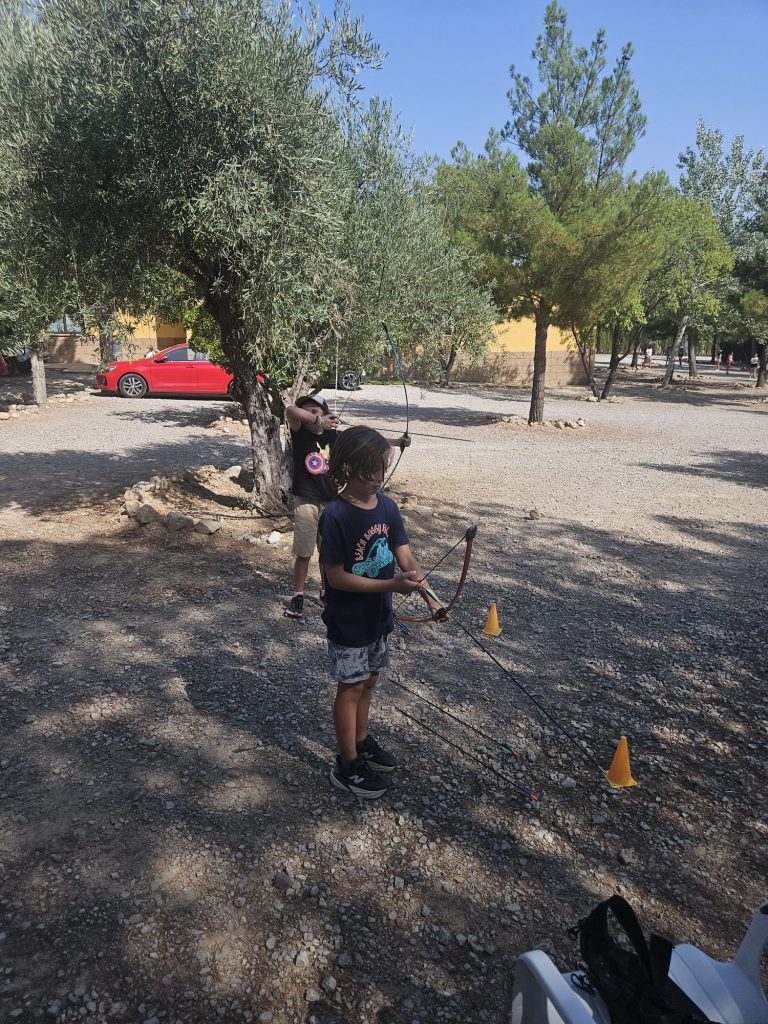 Two children practice archery outdoors among olive trees and gravel, with a yellow building and a red car in the background.