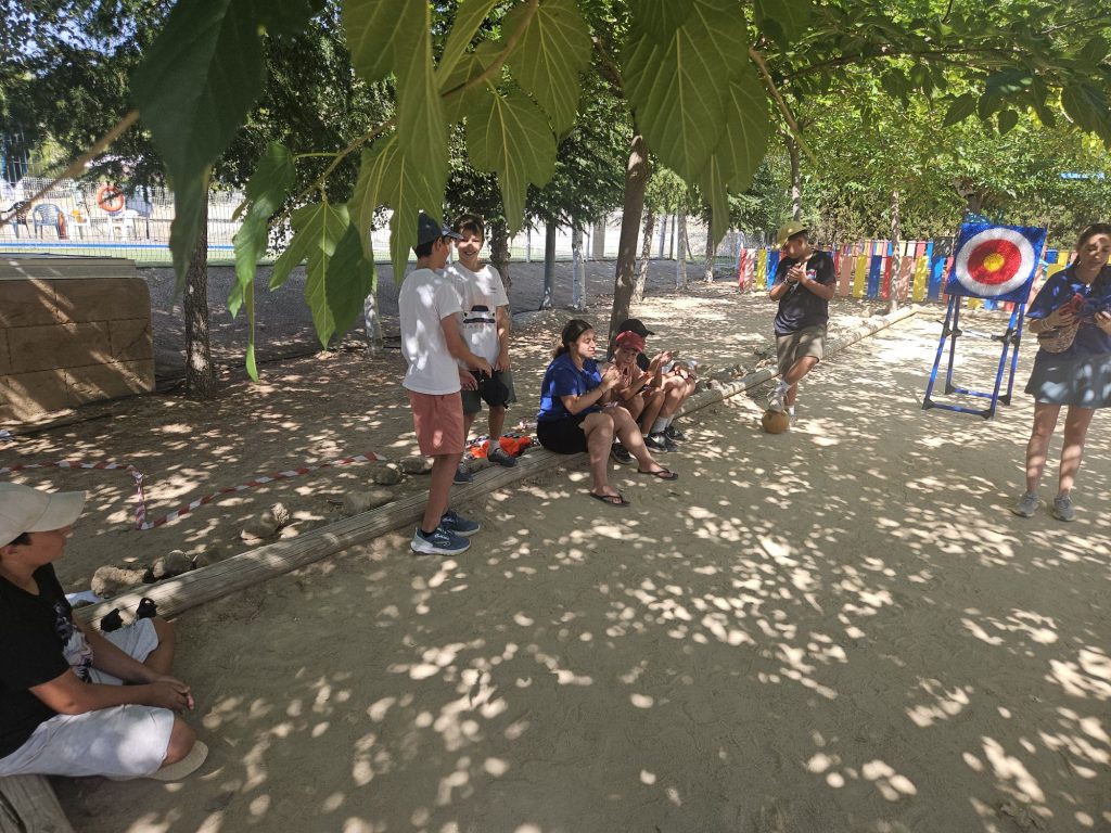 Group of kids and adults outdoors under trees, sitting and standing near a colorful archery target and fencing in the background.