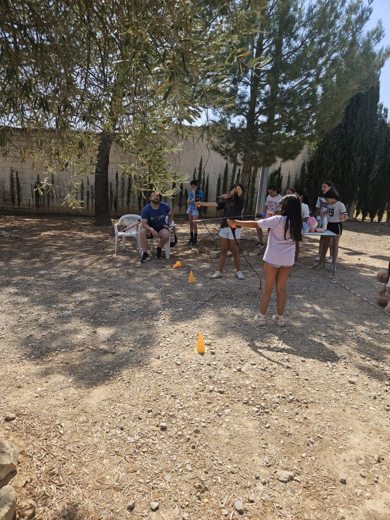 Group of kids learning archery outdoors, aiming arrows while a man sits on a white plastic chair nearby under a tree.