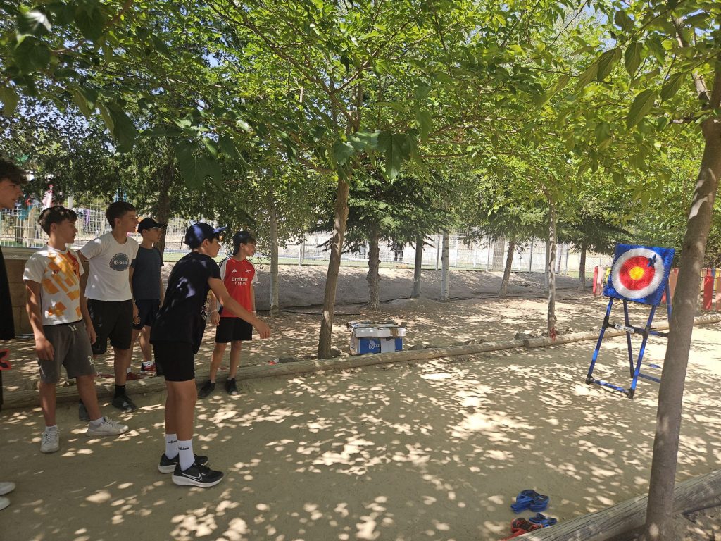 Group of boys standing under trees in an outdoor area, facing a bright archery target on a stand to the right.