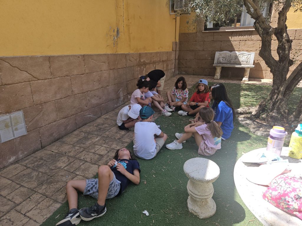 Group of children sitting in a circle on a courtyard, near a yellow wall and a tree.