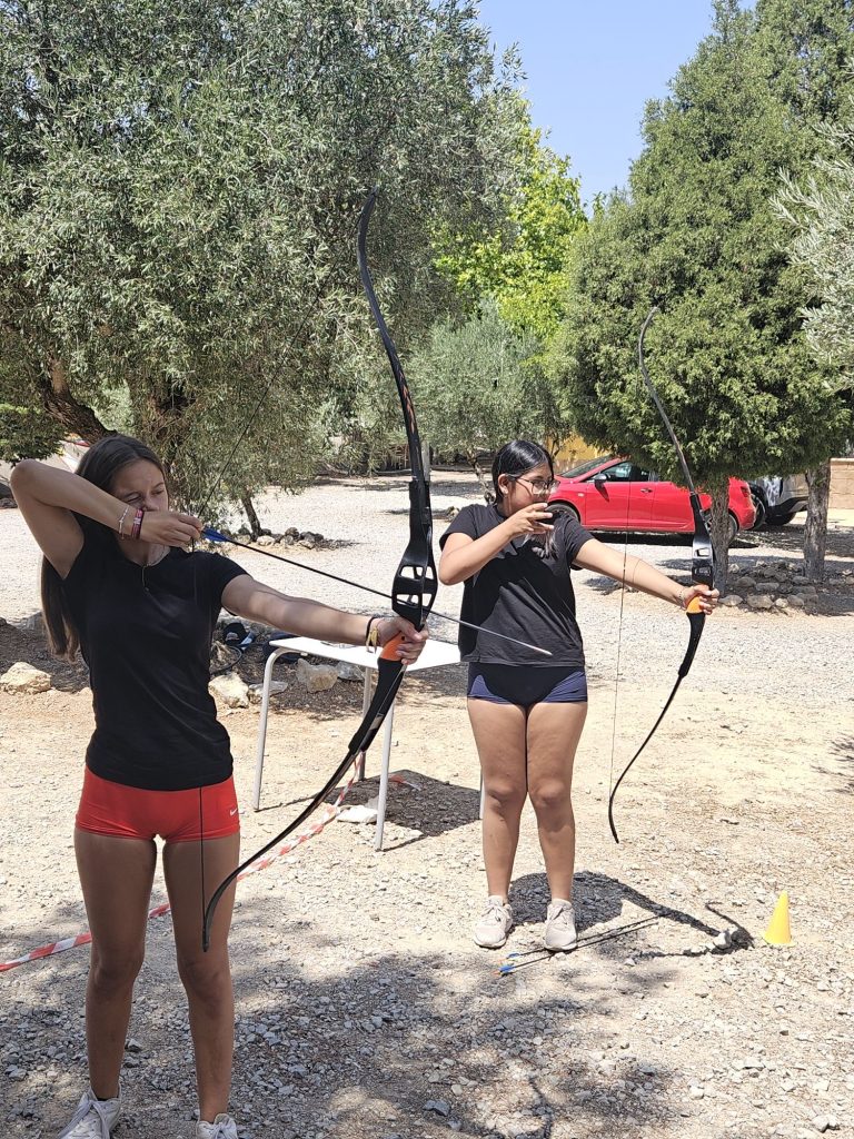 Two young women practice archery outdoors, both drawing bows and aiming off to the left, with trees and a red car in the background.