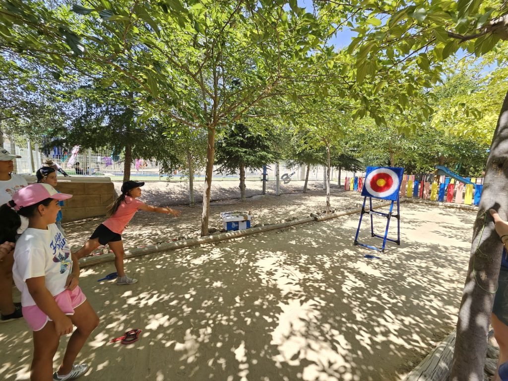Children in a shade-dappled park watching someone throw a small item toward a colorful target on a stand near a fence.