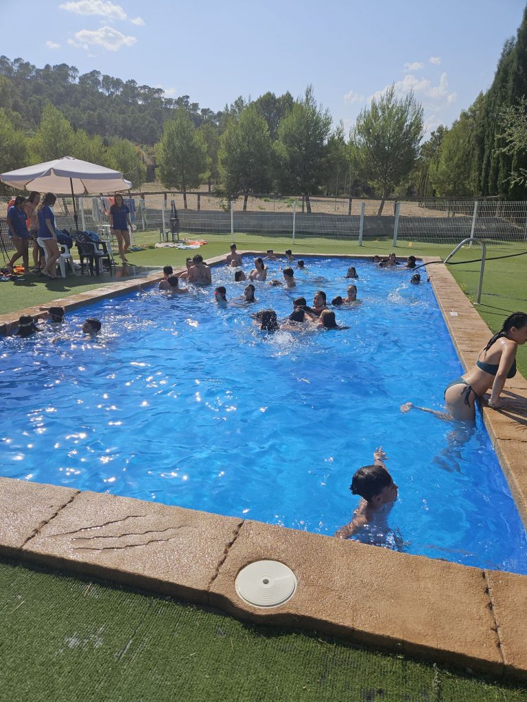 Outdoor swimming pool crowded with people, mostly kids, enjoying a sunny day under a large white umbrella by a grassy area and fence.