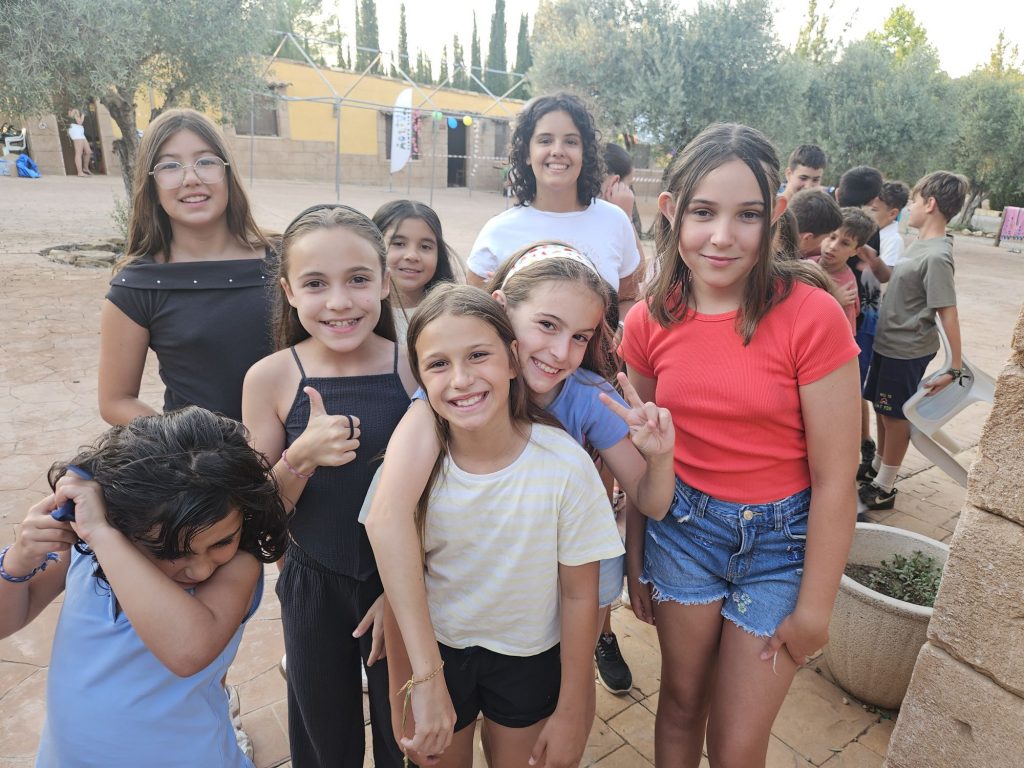 Group of smiling children posing together outdoors in a sunny courtyard, some making peace signs and thumbs up.