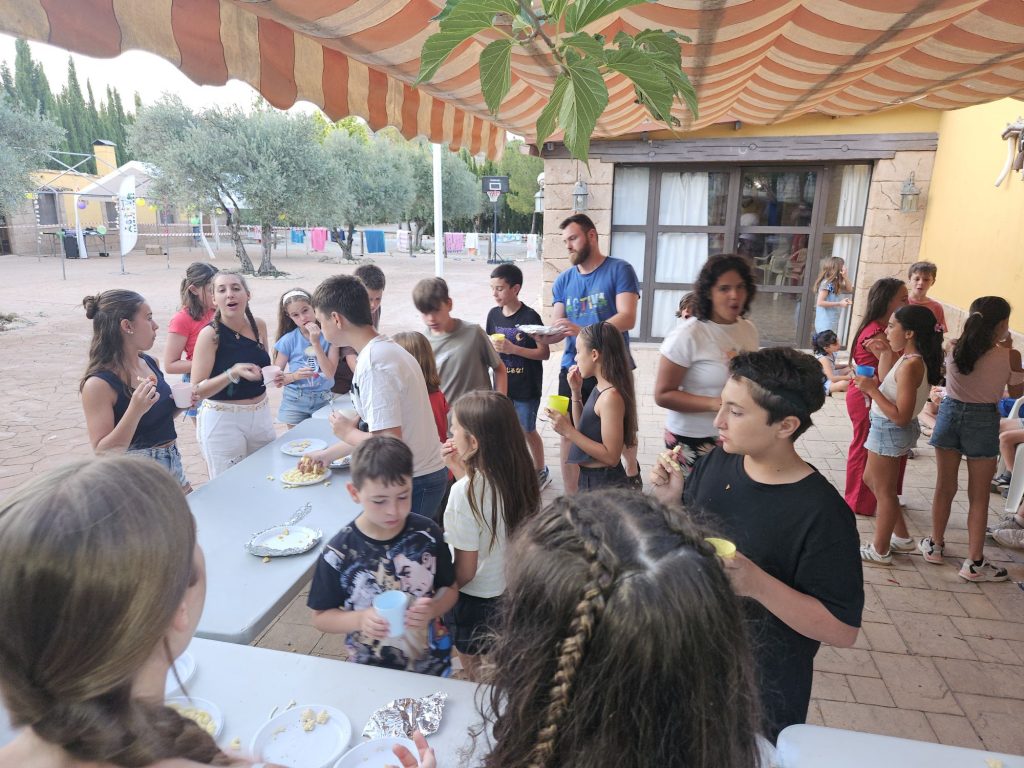 Group of kids and a few adults gathered around a long table with snacks and drinks at an outdoor party under a striped canopy.