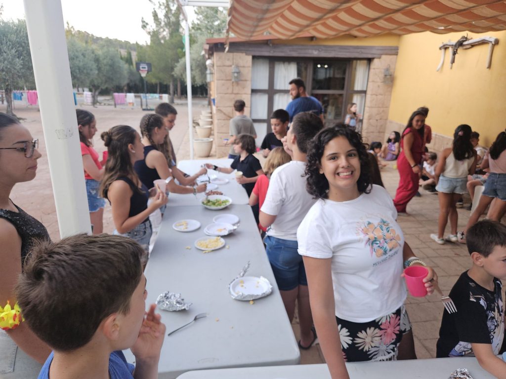 Group of children at an outdoor buffet under a striped canopy, serving themselves food from a long table next to a yellow building.