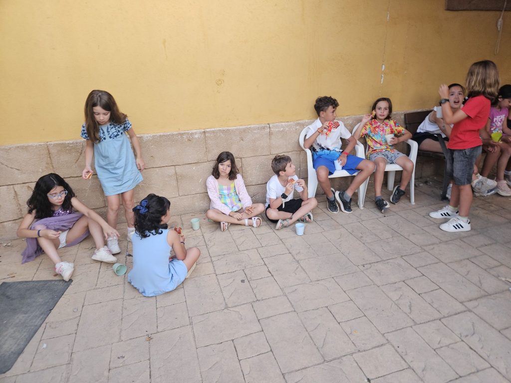 Group of children sitting and standing along a yellow wall, eating snacks at an outdoor gathering.