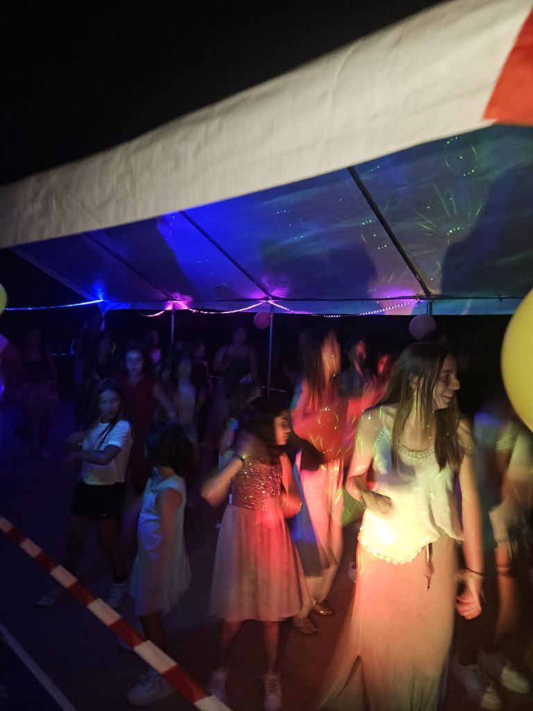 People wearing formal dresses dancing under a illuminated tent with colorful lights and balloons at a night party.