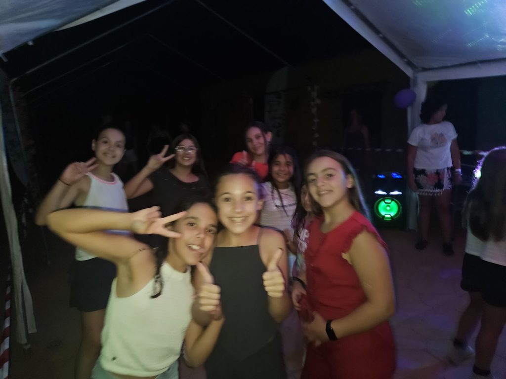 Group of smiling children posing for a photo at a night party under a tent, giving thumbs up and peace signs.
