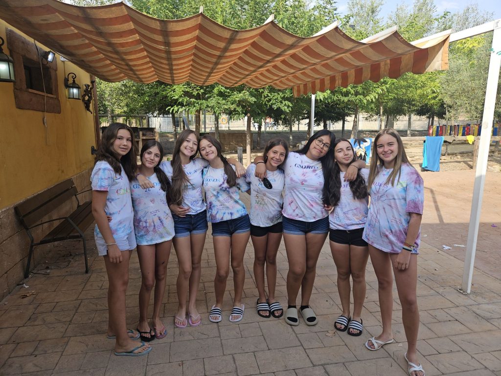 Group of smiling teenage girls in tie-dye T-shirts posing together outdoors under a striped fabric canopy.