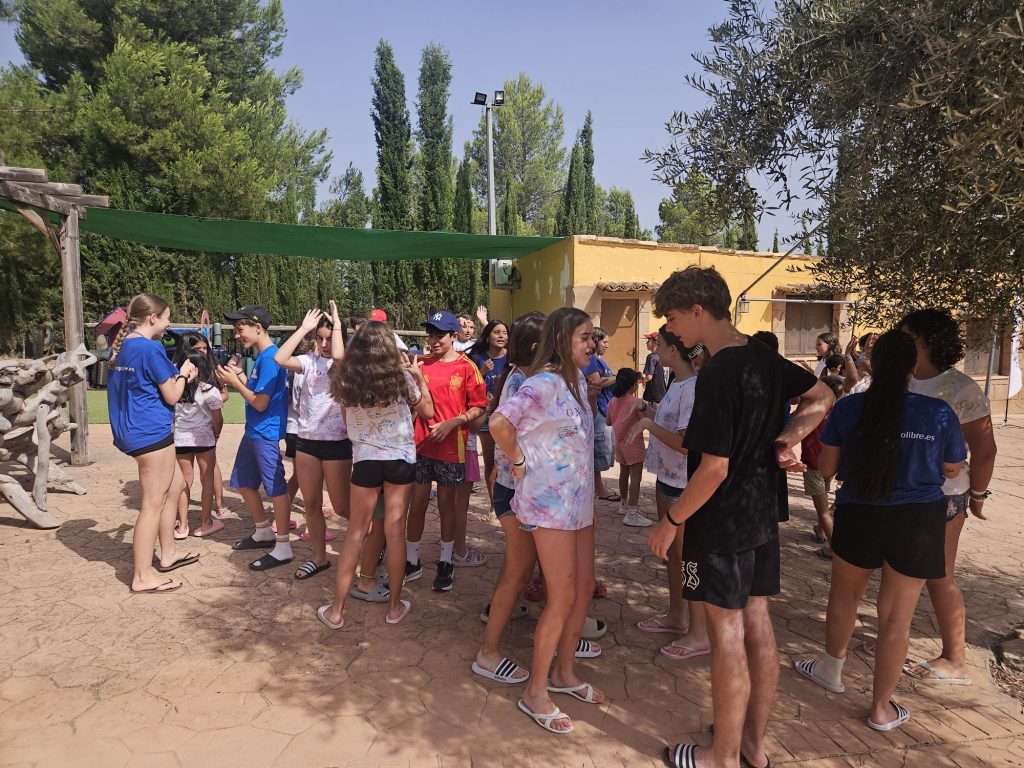 Group of smiling kids and teens in colorful shirts gather outdoors at a sunny camp, near a yellow building and green shade.