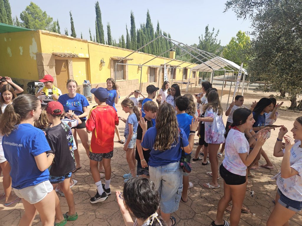 Group of children in blue shirts gathered outdoors in a sunny courtyard beside a yellow building, some wearing caps and interacting with each other.