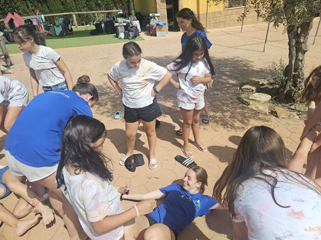 Children in a circle outdoors, holding hands and smiling during a group game or activity on a sunny day.