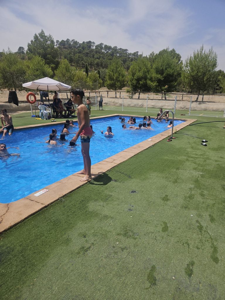 Children swimming in a bright outdoor pool with a boy standing at the edge by the poolside, under a white umbrella.