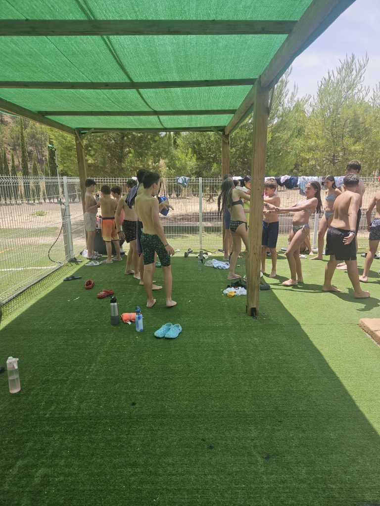 Group of children in swimsuits lined up under a green shade canopy at an outdoor pool area, towels and shoes scattered on artificial grass.
